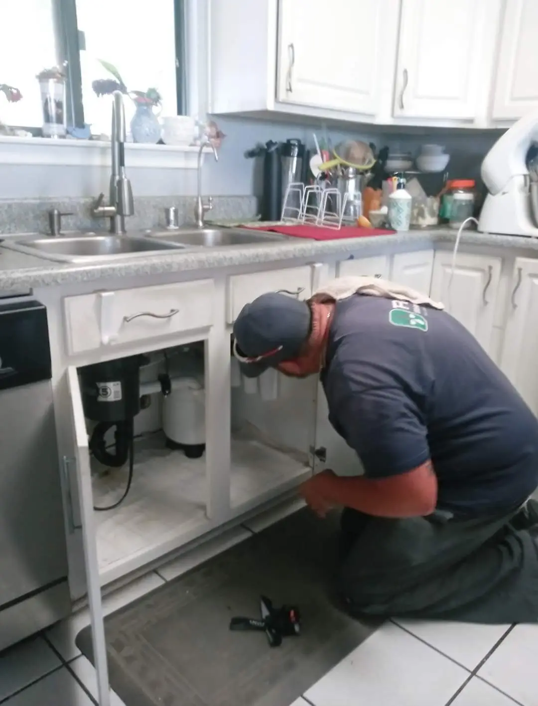 Service John A technician crouches beside a kitchen sink, inspecting pipes under the cabinet while tools are laid out on the floor.